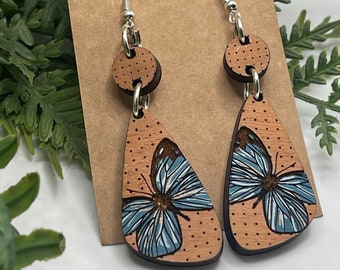 Puede incluir: Archivo de corte SVG para un par de pendientes de madera con un diseño de mariposa azul sobre un fondo marrón. Los pendientes están sobre una superficie marrón con follaje verde en el fondo. El texto "SVG ONLY NO ITEM WILL BE SENT" es visible en la imagen.