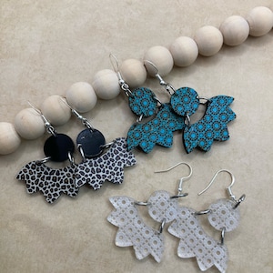 May include: Three pairs of dangle earrings with different designs. The first pair is black and white with a leopard print. The second pair is turquoise blue with a geometric pattern. The third pair is white with a lace-like pattern.