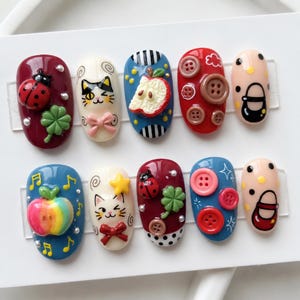 Può includere: Un set di dieci unghie finte con decorazioni 3D tra cui coccinelle, quadrifogli, gatti con fiocchi, fette di mela, bottoni colorati e scarpe, realizzate in rosso, blu, crema e rosa con vari motivi decorativi e dettagli grafici.