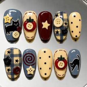 May include: A set of ten press-on nails featuring a variety of designs including black cat silhouettes, red tomatoes with button centers, cream-colored stars with button centers, blue and cream plaid patterns, and black polka dots on cream bases.