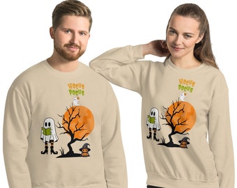 Puede incluir: Sudaderas beige con un diseño de Halloween. El diseño presenta un fantasma leyendo un libro, un árbol, una luna naranja y una calabaza con un sombrero de bruja. Las palabras "Hocus Pocus" están impresas encima del diseño en naranja y verde.