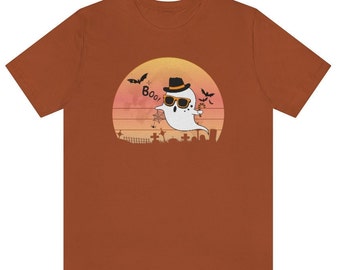 May include: A rust-colored t-shirt featuring a Halloween-themed graphic. The design includes a cartoon ghost wearing sunglasses and a hat, with the word "Boo!" in a speech bubble. Bats and a graveyard scene complete the design.