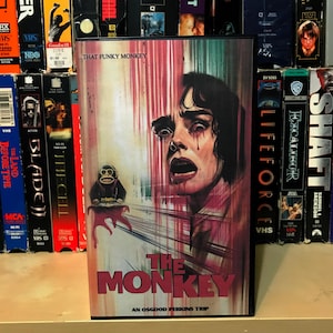 May include: A VHS tape cover for the horror film "The Monkey", featuring a red and white background with a close-up of a woman's face with blood dripping down her cheeks. The title "The Monkey" is in large, bold letters at the bottom of the cover. The text "An Osgood Perkins Trip" is below the title.