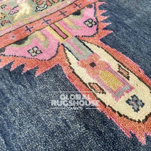 Op de afbeelding: Close-up van een deel van een tapijt met een marineblauwe achtergrond en een gedetailleerd, kleurrijk ontwerp. Het ontwerp heeft roze, oranje, beige en grijze geometrische patronen. Het tapijt heeft een getextureerde uitstraling.