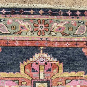 Op de afbeelding: Close-up van een gedeelte van een patroonkleed. Het kleed heeft een marineblauw veld met een centraal ontwerp in roze, geel en oranje. De rand bevat een grijsgroene band met bloemen- en paisley-motieven en een roze en oranje streep.