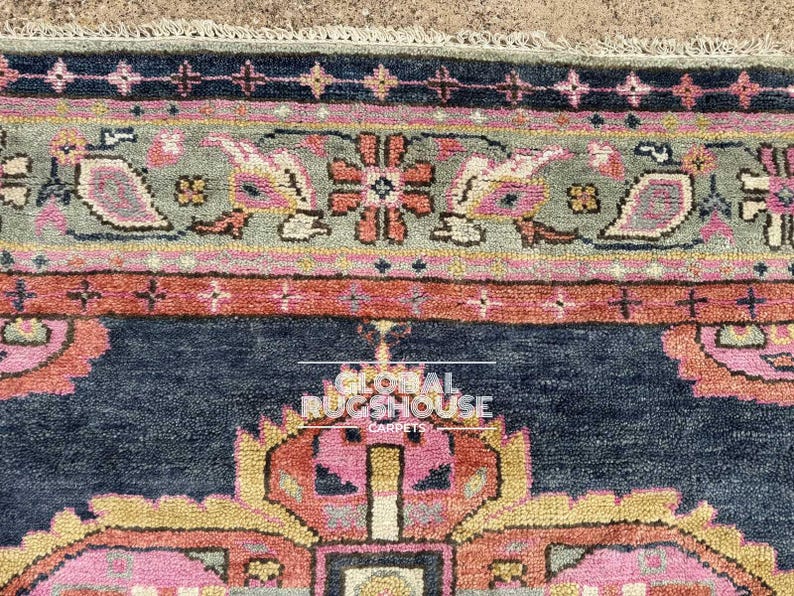 Op de afbeelding: Close-up van een gedeelte van een patroonkleed. Het kleed heeft een marineblauw veld met een centraal ontwerp in roze, geel en oranje. De rand bevat een grijsgroene band met bloemen- en paisley-motieven en een roze en oranje streep.