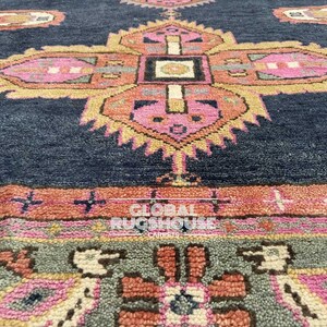 Op de afbeelding: Close-up van een marineblauw tapijt met een gedetailleerd, geometrisch patroon. Het ontwerp heeft roze, oranje en gele bloemmotieven. Het tapijt heeft een getextureerd uiterlijk, met de woorden "GLOBAL RUGSHOUSE CARPETS" zichtbaar.