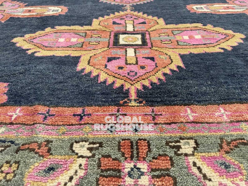 Op de afbeelding: Close-up van een marineblauw tapijt met een gedetailleerd, geometrisch patroon. Het ontwerp heeft roze, oranje en gele bloemmotieven. Het tapijt heeft een getextureerd uiterlijk, met de woorden "GLOBAL RUGSHOUSE CARPETS" zichtbaar.