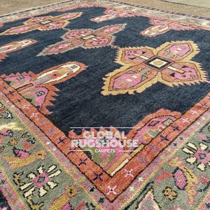 Op de afbeelding: Een groot, vierkant tapijt met een donkerblauwe achtergrond en ingewikkelde patronen in roze, oranje en geel. Het tapijt heeft een centraal ontwerp met herhaalde bloemen- of geometrische motieven en een gedetailleerde rand.