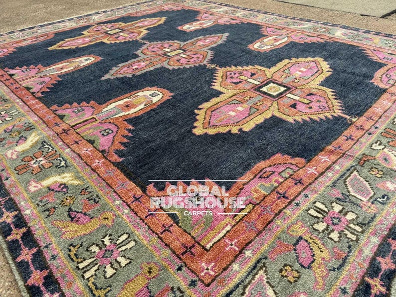 Op de afbeelding: Een groot, vierkant tapijt met een donkerblauwe achtergrond en ingewikkelde patronen in roze, oranje en geel. Het tapijt heeft een centraal ontwerp met herhaalde bloemen- of geometrische motieven en een gedetailleerde rand.