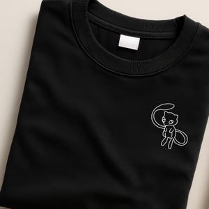 Könnte beinhalten: Schwarzes Rundhals-T-Shirt mit einer weißen Umrissgrafik eines cartoonartigen, katzenartigen Wesens auf der Vorderseite. Das Shirt ist sauber gefaltet und zeigt den Kragen und ein kleines Etikett. Das Design ist schlicht und sauber.