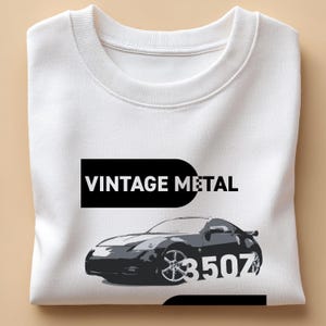 Puede incluir: Sudadera blanca con un gráfico de un coche deportivo gris y el texto "VINTAGE METAL" encima. El coche tiene el número "350Z" en el lateral. La sudadera está doblada cuidadosamente.
