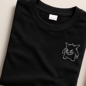 May include: Black crew neck sweatshirt with a white outline graphic of a cartoon monster on the front. The sweatshirt is folded neatly, showcasing the design. The design is a stylized, grinning monster.