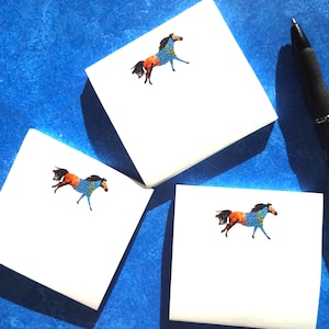 Könnte beinhalten: Drei quadratische weiße Haftnotizen, jede mit einer kleinen, farbenfrohen Illustration eines laufenden Pferdes mit orangefarbenen, blauen und schwarzen Mustern. Ein schwarzer Kugelschreiber liegt rechts neben den Notizen auf einer blauen Fläche.