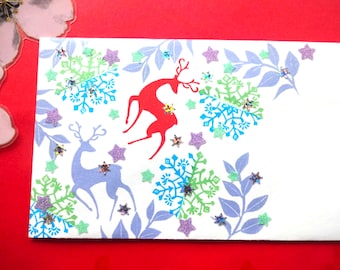 Könnte beinhalten: Eine weiße rechteckige Grußkarte mit einer roten Hirschsilhouette, einer hellvioletten Hirschsilhouette sowie blauen und grünen Schneeflockenmustern. Das Design ist mit kleinen violetten und grünen sternförmigen Glitzerpartikeln und hellvioletten Blattmotiven verziert.