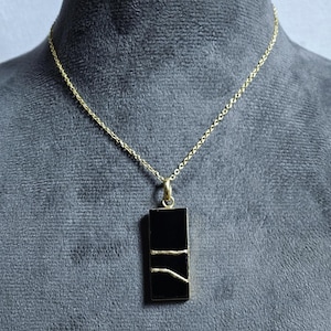 Könnte beinhalten: Eine goldene Kettenkette mit einem schwarzen rechteckigen Anhänger mit dünnen goldenen Linien.