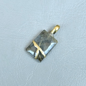 May include: A rectangular pendant with a gold-colored bail and a gold-colored accent. The pendant is a mix of gray and blue-green with a gold-colored accent running across the center. The pendant is approximately 1 inch long.
