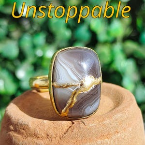 Può includere: Un anello d'oro con una grande pietra rettangolare grigia e bianca. La pietra ha un accento dorato. La parola "Unstoppable" è scritta in giallo sopra l'anello.