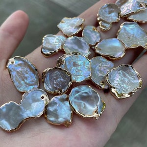 Pode incluir: Uma coleção de pingentes de concha de pérola barroca iridescente branca e azul com detalhes metálicos dourados. As conchas têm uma forma irregular e um aspeto natural e orgânico.