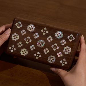 May include: A rectangular wooden box featuring a dark brown finish and an inlaid pattern of iridescent mother-of-pearl floral and heart-shaped motifs across the top surface. The box has rounded edges and a smooth, polished wood grain texture.