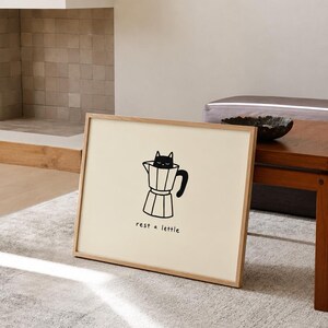 Puede incluir: Una lámina de arte enmarcada que presenta un dibujo de línea negra de un gato sentado dentro de una cafetera moka. La ilustración está centrada sobre un fondo color crema con el texto en minúsculas "rest a lettle" impreso bajo la cafetera.