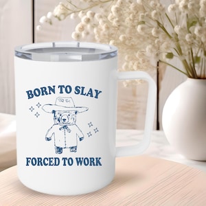 Pode incluir: Caneca de cerâmica branca com um design azul. A caneca tem uma alça e diz "Born to Slay" com um urso de desenho animado usando um chapéu de cowboy e "Forced to Work" embaixo.