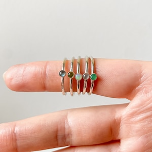 Könnte beinhalten: Fünf Silberringe mit verschiedenen farbigen Edelsteinen. Die Ringe sind übereinander auf einem Finger gestapelt. Die Edelsteine sind blau, grün, weiß, grau und grün.