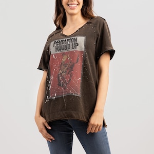 May include: A brown t-shirt with a distressed look and a graphic print of a cowboy riding a bucking bronco. The graphic has the text "PENDLETON ROUND UP" and "PENDLETON OREGON".