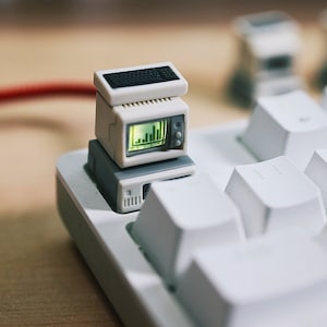 Puede incluir: Un conjunto de ordenador en miniatura, que incluye un monitor, un teclado y una CPU, está colocado sobre una tecla blanca del teclado. El monitor muestra un gráfico de barras verde. El conjunto es de color gris claro.