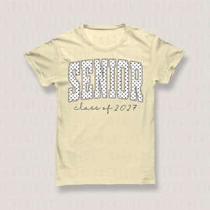 Puede incluir: Camiseta amarilla clara con la palabra "SENIOR" en letras grandes blancas con lunares. Debajo, el texto "class of 2027" est&aacute; escrito en negro fino. La camiseta tiene mangas cortas y cuello redondo.