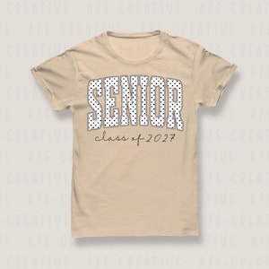 Puede incluir: Una camiseta beige con la palabra "SENIOR" en letras grandes con lunares blancos. Debajo, el texto "class of 2027" est&aacute; escrito en negro. La camiseta tiene mangas cortas y cuello redondo.
