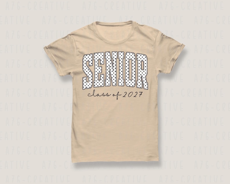 Puede incluir: Una camiseta beige con la palabra "SENIOR" en letras grandes con lunares blancos. Debajo, el texto "class of 2027" est&aacute; escrito en negro. La camiseta tiene mangas cortas y cuello redondo.