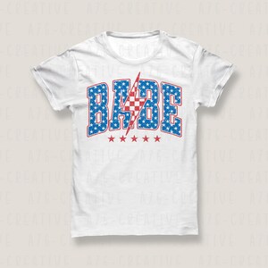 May include: White t-shirt with a red, white, and blue graphic that says "BABE" with a lightning bolt in the middle. The graphic is made up of stars and stripes.