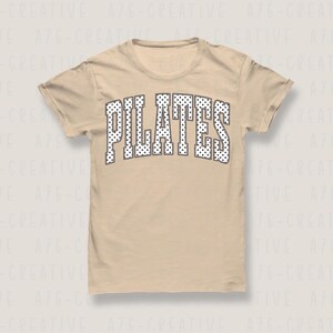 May include: A beige short-sleeve t-shirt featuring the word PILATES printed in a large, arched, collegiate-style font. The letters are white with a black outline and are filled with a repeating pattern of small black polka dots.