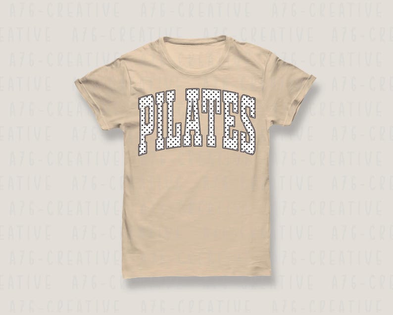 May include: A beige short-sleeve t-shirt featuring the word PILATES printed in a large, arched, collegiate-style font. The letters are white with a black outline and are filled with a repeating pattern of small black polka dots.