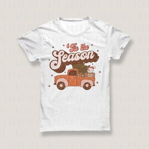Op de afbeelding: Wit T-shirt met een retro ontwerp. De tekst "'Tis the Season" staat boven een oranje pick-up truck die een kerstboom en een sneeuwpop vervoert. Kleine sterren zijn verspreid rond de tekst en de truck.