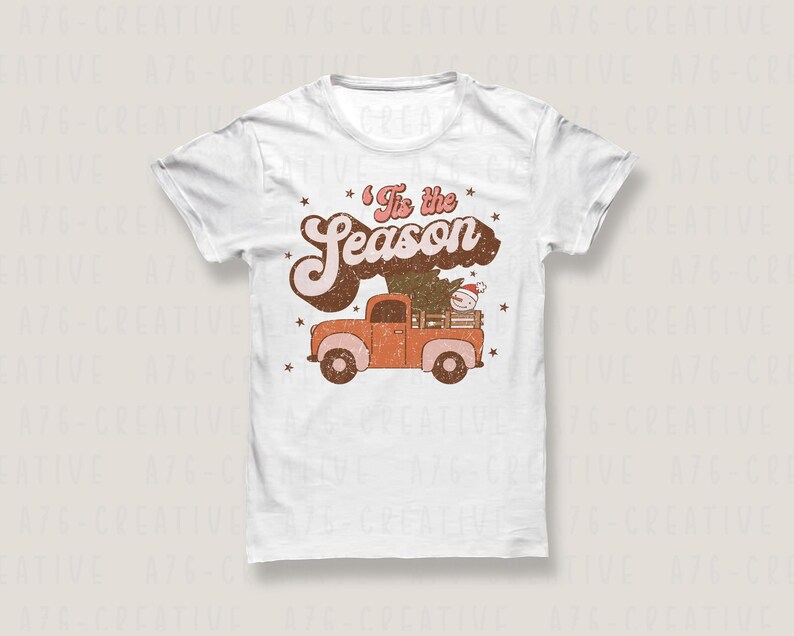 Op de afbeelding: Wit T-shirt met een retro ontwerp. De tekst "'Tis the Season" staat boven een oranje pick-up truck die een kerstboom en een sneeuwpop vervoert. Kleine sterren zijn verspreid rond de tekst en de truck.