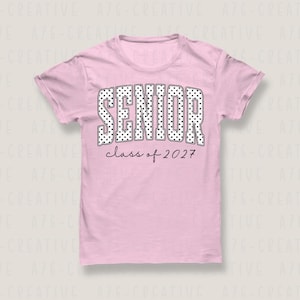 Puede incluir: Camiseta rosa claro con la palabra "SENIOR" en letras grandes blancas con lunares. Debajo, el texto "class of 2027" en fuente fina negra. La camiseta es de cuello redondo, ideal para eventos escolares.