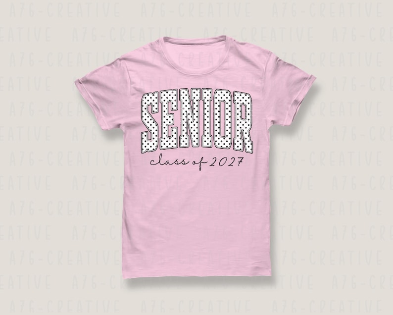 Puede incluir: Camiseta rosa claro con la palabra "SENIOR" en letras grandes blancas con lunares. Debajo, el texto "class of 2027" en fuente fina negra. La camiseta es de cuello redondo, ideal para eventos escolares.