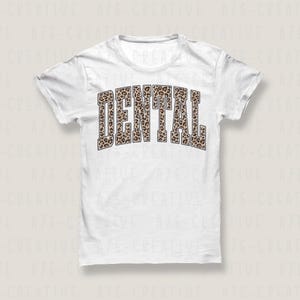 May include: A white t-shirt with the word "DENTAL" printed across the front in a bold, arched font. The letters are filled with a brown and tan leopard print pattern, creating a striking contrast against the white fabric.