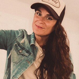 May include: A trucker hat with a brown brim and a cream-colored front panel. The front panel features the text "stay wild" in a cursive font. The hat is worn with a denim jacket.