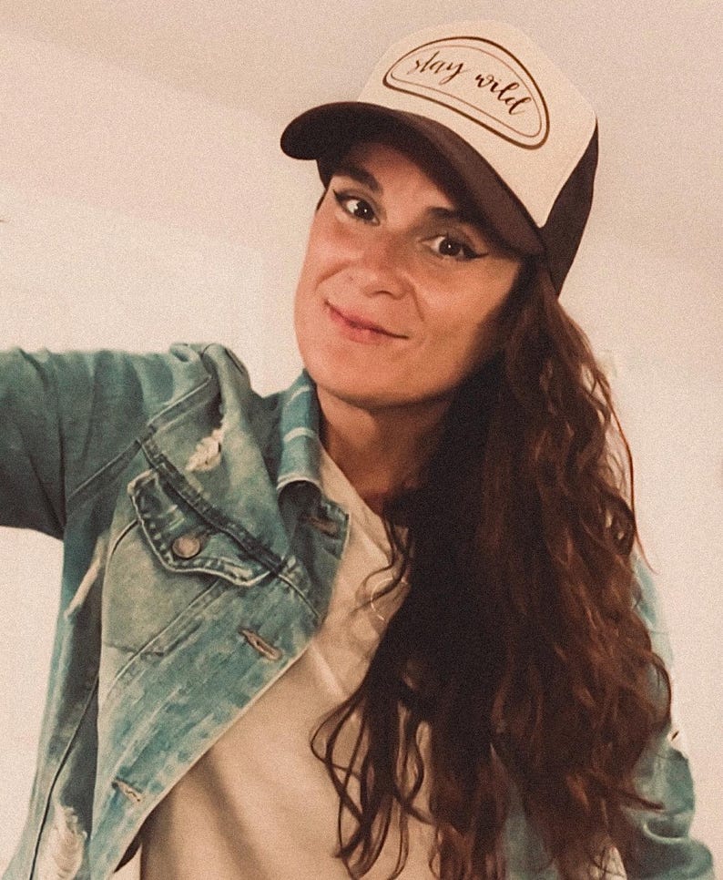 May include: A trucker hat with a brown brim and a cream-colored front panel. The front panel features the text "stay wild" in a cursive font. The hat is worn with a denim jacket.
