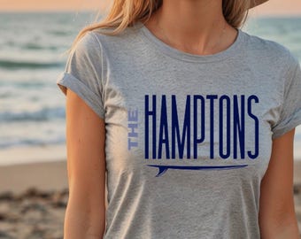 May include: A heather gray short-sleeved t-shirt featuring the dark blue text THE HAMPTONS printed on the chest. Below the text is a dark blue graphic of a surfboard. The shirt has a crew neckline and rolled sleeves, providing a casual appearance.