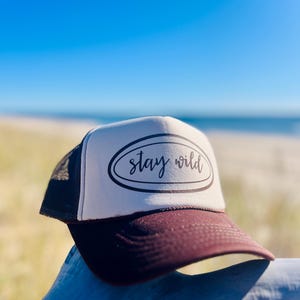 May include: A trucker hat with a white front panel and a dark burgundy brim. The hat features the text "stay wild" in a black script within a black oval. The hat is set against a backdrop of a beach and blue sky.