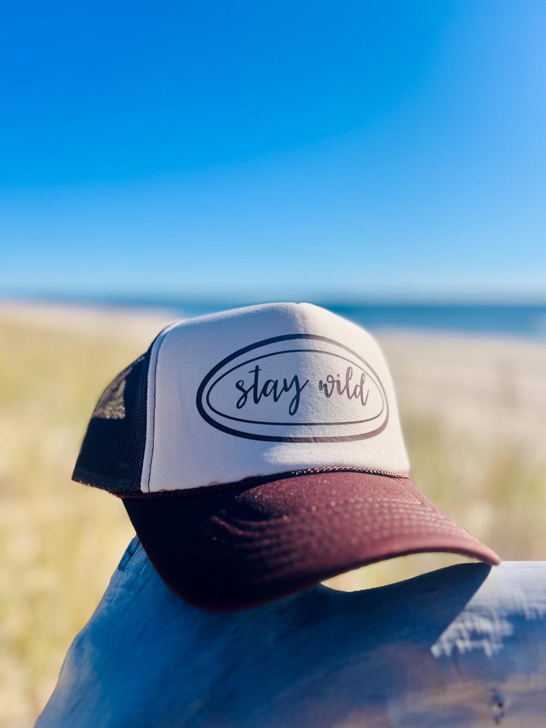 May include: A trucker hat with a white front panel and a dark burgundy brim. The hat features the text "stay wild" in a black script within a black oval. The hat is set against a backdrop of a beach and blue sky.
