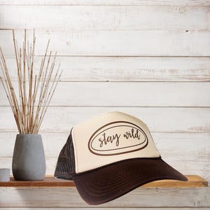 May include: A trucker hat with a brown brim and mesh back, and a cream-colored front panel. The front panel features the words "stay wild" in a brown oval design. The hat is displayed on a wooden shelf against a white wood-paneled background.