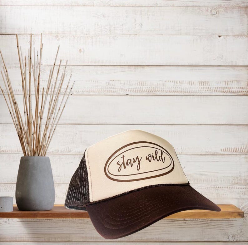 May include: A trucker hat with a brown brim and mesh back, and a cream-colored front panel. The front panel features the words "stay wild" in a brown oval design. The hat is displayed on a wooden shelf against a white wood-paneled background.