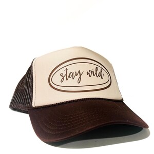 May include: A brown and tan trucker hat with the text "Stay Wild" in a brown oval. The hat has a brown brim and mesh backing. The hat is a casual accessory.
