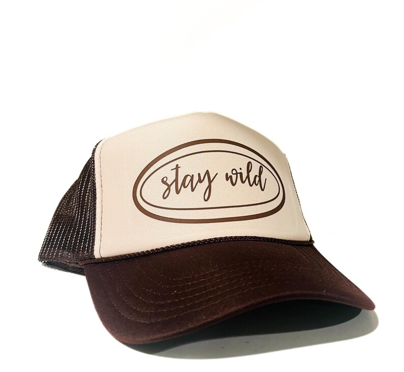 May include: A brown and tan trucker hat with the text "Stay Wild" in a brown oval. The hat has a brown brim and mesh backing. The hat is a casual accessory.