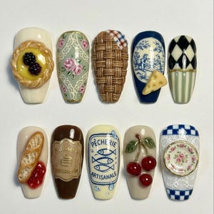 May include: A set of ten artificial nails with various designs. The designs include a tart with berries, floral patterns, woven basket, cheese, a blue and white scene, a black and white diamond pattern, a baguette, a patisserie label, fish, cherries, and a floral plate.